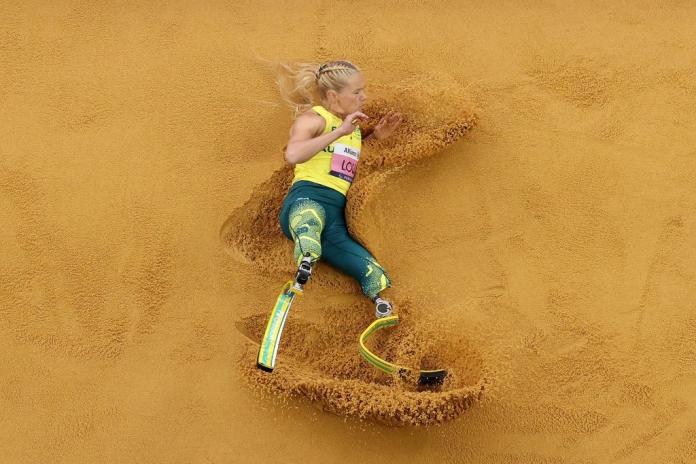 A photo taken from above shows a female athlete wearing running blades landing in the sand pit